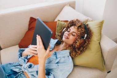 Mutlu Kadın, tablet, bilgisayar ve kulaklık tutuyor, müzik ve rahat bir evde kanepede dinleniyor.