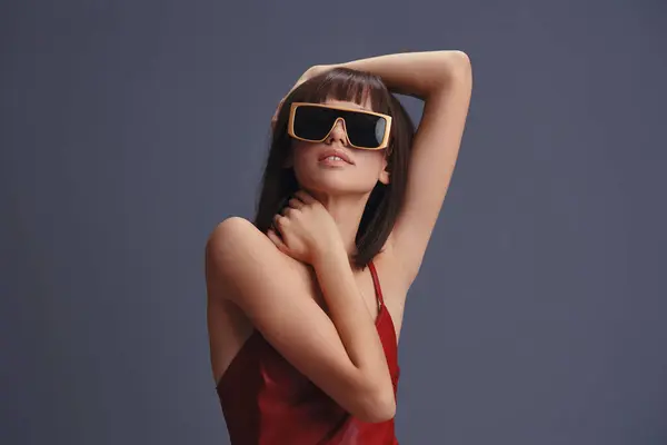 stock image A woman in a red dress and sunglasses stands on a gray background, posing with her hands on her hips and her head tilted to the side, exuding confidence and elegance