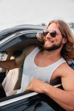 Kolsuz tişörtlü neşeli bir adam arabanın camından sarkmış, parlak bir gülümsemeyle yaz melteminin tadını çıkarıyor.
