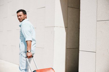 Gündelik kıyafetli genç bir adam kırmızı bir bavul çeker, beyaz duvarlı modern bir binanın yanında yürürken arkasına bakar..