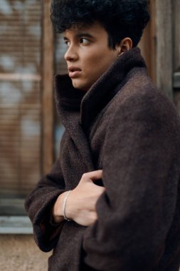 Kahverengi ceketli modacı, tuğla duvara yaslanmış, elleri kalçasında ve kolları göğsünde çaprazlanmış..