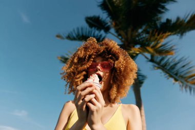 Kıvırcık saçlı neşeli genç bir kadın açık mavi gökyüzüne karşı palmiye ağaçlarıyla çevrili bir dondurma külahını tutuyor.
