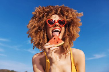 Kıvırcık saçlı neşeli kadın karpuz yiyor, neşe saçıyor canlı mavi gökyüzüne ve güneşli arkaplana