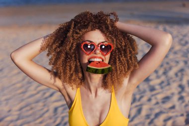 Neşeli genç bir kadın kumlu sahilde poz veriyor, elinde bir dilim karpuz, neşeli bir gülümsemeyle, yaz havasını yansıtıyor.