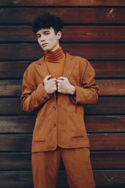 Şık kahverengi takım elbiseli genç bir adam koyu ahşap bir duvarın önünde duruyor, modern moda tarzını sergiliyor.