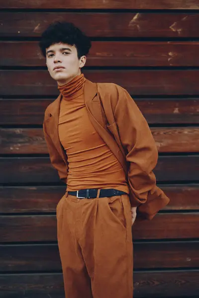 Stock image A fashionable young man wearing a fitted orange turtleneck and loose pants posing near a rustic wooden wall