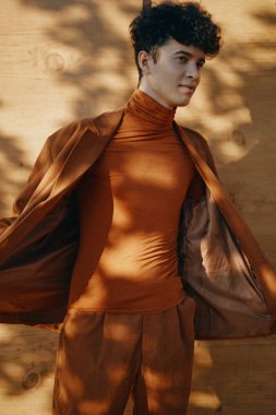 Havalı bir erkek model şık kahverengi balıkçı yaka ve takım elbisesini açık havada sergiliyor. Yumuşak gölgeli ağaçlar havayı güzelleştiriyor.