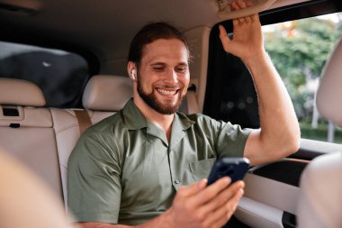 Yeşil tişörtlü, gülümseyen bir adam, lüks bir arabanın içinde, hafif ışıklandırmayla çevrili bir şekilde otururken telefonla konuşmaktan zevk alır.