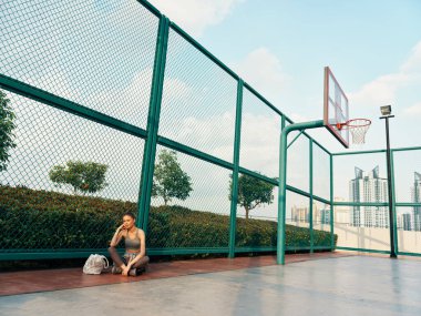 Bir kadın basketbol sahasının zemininde oturuyor, düşünceli görünüyor, etrafı çitlerle çevrili ve kentsel manzaralı.