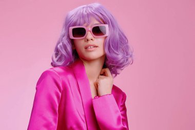 Mor saçlı, güneş gözlüğü takan ve pembe takım elbiseli bir kadın pembe bir arka plana karşı duruyor. Parlak saç rengi ve uyumlu kıyafetiyle cesur ve şık bir moda ifadesi sergiliyor.