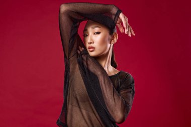 Asyalı manken, siyah bluzlu, kırmızı arka planda poz veriyor, elleri kalçada, başı yana dönük..