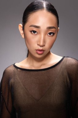 Gri bir arkaplan önünde poz veren katıksız bluzlu moda Asyalı kadın. Şık Asyalı bir kadın kameraya özgüvenle poz veriyor..