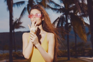 Sarı elbiseli neşeli genç bir kadın güneş gözlüğü takıyor, tropik palmiye ağaçları ve mavi gökyüzüne karşı dondurmanın tadını çıkarıyor.
