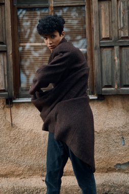 Moda: kahverengi ceketli ve siyah pantolonlu genç bir adam bir pencerenin önünde duruyor, elleri kalçasında, başı yana yatık..