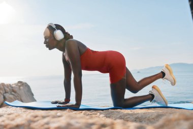 Formda bir kadın, güzel bir deniz manzarasının arka planında mavi bir yoga minderi üzerinde egzersiz yapıyor.