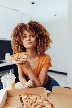 Kıvırcık saçlı kadın pizza kutusunun önünde bir dilim pizza tutuyor. Lezzetli bir İtalyan yemeğinin tadını çıkarıyor.