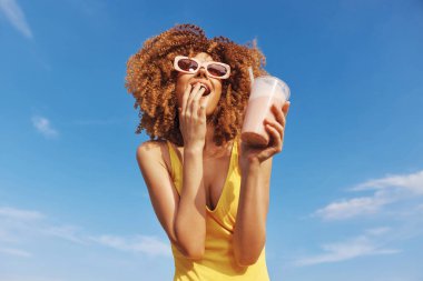 Kıvırcık saçlı neşeli bir kadın canlı mavi gökyüzüne karşı içki içiyor, şık güneş gözlükleri takıyor ve parlak bir mayo giyiyor.