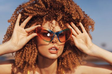 Modayı takip eden bir kadın, açık mavi gökyüzü ve rahat bir atmosfere sahip güneşli bir arka planda canlı güneş gözlükleri sergiliyor.