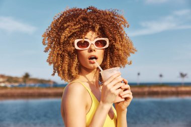 Kıvırcık saçlı, güneş gözlüğü takan şık bir kadın ferahlatıcı bir içecek tutuyor, açık mavi gökyüzüne ve okyanus zeminine karşı.