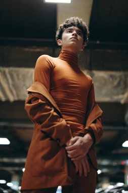 Zarif bir genç adam, bir otoparkın endüstriyel estetiğiyle çevrili parlak kahverengi bir bluz ve ceketle poz veriyor. Kendine güveni modern sokak tarzını vurguluyor.