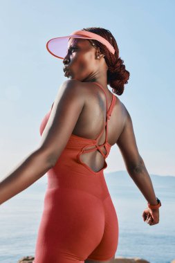 Canlı kırmızı kıyafetli atletik bir kadın mavi bir okyanus zeminine karşı duruyor, enerji ve pozitiflik saçıyor.