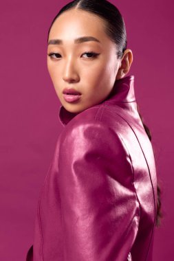 Mor deri ceketli şık Asyalı esmer, bir moda fotoşopu için pembe arka planda poz veriyor..