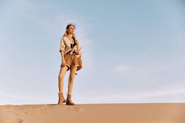 Çölün ortasında, açık mavi gökyüzünün altında, kum tepesinde duran bir kadın.
