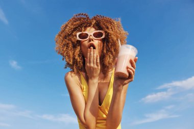 Güneş gözlüklü, parlak sarı elbiseli, açık havada serinletici bir meyve püresinin tadını çıkaran sürpriz bir genç kadın.