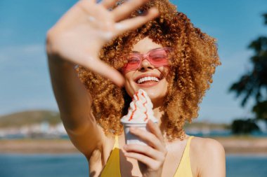 Kıvırcık saçlı, güneş gözlüğü takan neşeli bir kadın dondurma tutuyor ve kumsalda güneşli bir yaz gününün tadını çıkarıyor.
