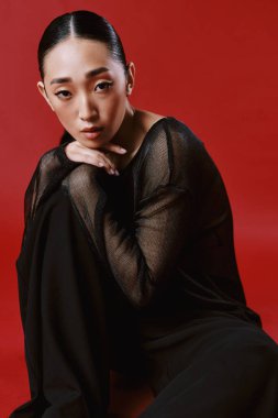 Siyah elbiseli Asyalı kadın kırmızı arka planda oturuyor elleri dizlerinde ve çenesi göğsünde. moda portresi.