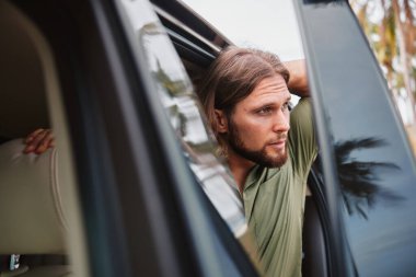 Uzun saçlı ve sakallı bir adam etrafı doğayla çevrili bir arabanın camından dikkatlice dışarı bakar.