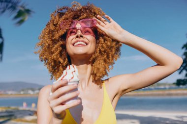 Sarı mayo giymiş, elinde dondurma tutan, okyanus kıyısında pembe güneş gözlüklü neşeli genç bir kadın.