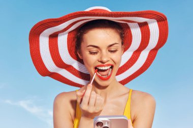 Geniş kenarlı şapkalı, açık havada güneşli bir günün tadını çıkarırken dudaklarını parlak rujla büyüten neşeli bir kadının görüntüsü.