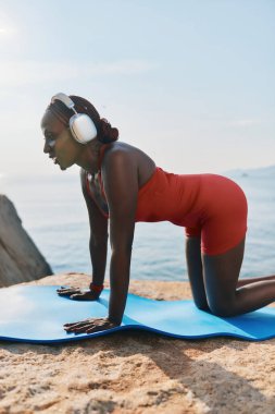 Kulaklık takan sağlıklı bir kadın okyanusun kenarındaki mavi bir mindere yoga pozu veriyor ve sabah güneşinin tadını çıkarıyor.