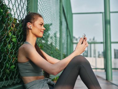 Bir kadın güneşli bir günde akıllı telefonundan hoşlanır, yeşil bir çite yaslanır, atletik kıyafetler giyer ve kablosuz kulaklık takar, rahat bir titreşim yayar.
