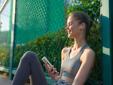 Kulaklık takan genç bir kadın dışarıda, elinde akıllı bir telefonla yeşil bir çitin karşısında otururken neşeli ifadesini vurguluyor.