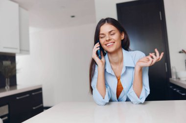 Mutfak masasında oturan kadın telefonla konuşuyor. İletişim ve teknoloji ile modern yaşam tarzı kavramı.