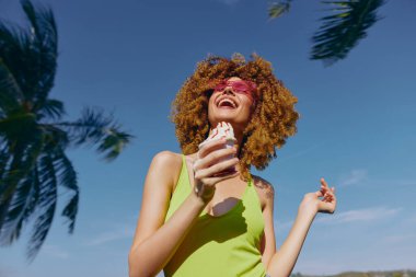 Kıvırcık saçlı bir kadın dondurma tutar, neşeyle gülümser arka planda parlak palmiye ağaçları ve parlak mavi bir gökyüzü vardır.