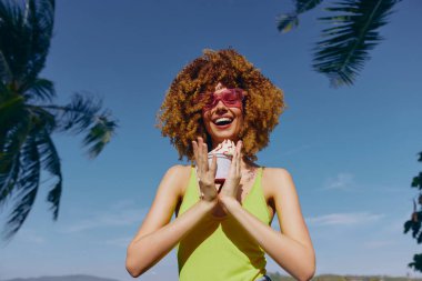 Parlak sarı bluzlu neşeli bir kadın, tropik palmiye yaprakları ve açık gökyüzü ile çevrili bir ziyafetin tadını çıkarıyor.