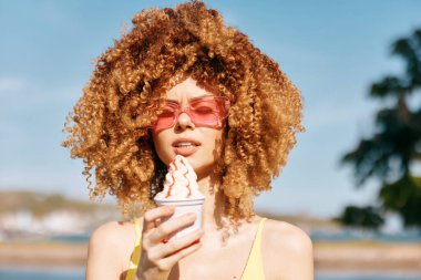 Canlı kıvırcık saçlı şık bir kadın elinde bir fincan dondurmayla açık mavi gökyüzüne ve yeşilliğe karşı.