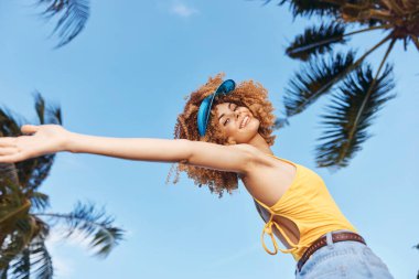Sarı kollu bluzlu ve kot şortlu neşeli genç bir kadın, parlak mavi gökyüzüne karşı, gür palmiye ağaçlarıyla çevrili, açık kollarıyla poz verir.