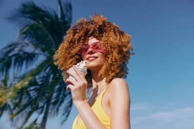 Kıvırcık saçlı ve güneş gözlüklü neşeli bir kadın açık mavi gökyüzüne ve palmiye ağacına karşı dondurmanın tadını çıkarıyor.