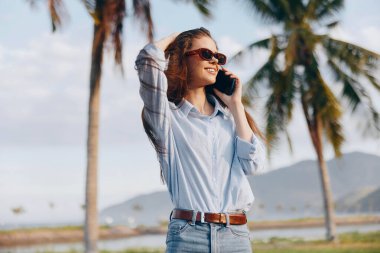 Güneş gözlüklü ve mavi gömlekli şık bir kadın cep telefonu, palmiye ağaçları ve sakin su arka planı, tropikal tatil atmosferi hakkında konuşuyor..
