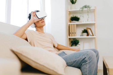 Genç adam, aydınlık, modern bir oturma odasındaki rahat bir koltukta otururken sanal gerçeklik deneyiminden zevk alıyor. Uzay, atmosferi aydınlatan minimalist dekorlar, sıcak tonlarda ve doğal ışık barındırıyor.