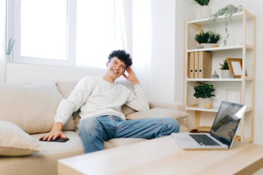 Genç bir adam rahat bir şekilde modern bir oturma odasında, rahat bir gülümsemeyle rahat bir şekilde oturur. Günlük kıyafetleri ve saksı bitkileri de dahil, güzel dekore edilmiş yeri, hoş bir karşılama havası yaratır.