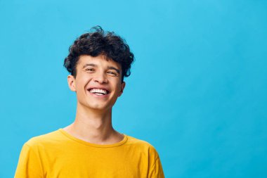 Kıvırcık saçlı neşeli genç adam sarı gömlek giyiyor, canlı mavi bir arka plana gülümsüyor bu görüntü mutluluğu, gençliği ve pozitifliği temsil ediyor, çeşitli projeler için ideal.