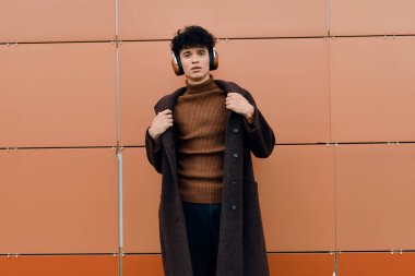 Kahverengi ceketli, kazaklı, siyah pantolonlu, kahverengi ayakkabılı, turuncu duvarın önünde duran, kulaklıklı şık bir genç..