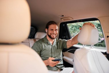 Neşeli bir adam arabanın arka koltuğunda eğleniyor, elinde akıllı bir telefon ve kablosuz kulaklık var açık renkli iç mekan sıcak bir hava yaratıyor.