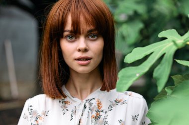 Doğadaki kızıl saçlı bir kadının portresi. Yeşilliklerin arasında duruyor ve izleyiciyle göz teması kuruyor.