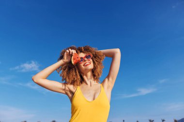Kıvırcık saçlı neşeli bir kadın karpuz şeklindeki güneş gözlükleriyle poz verir.